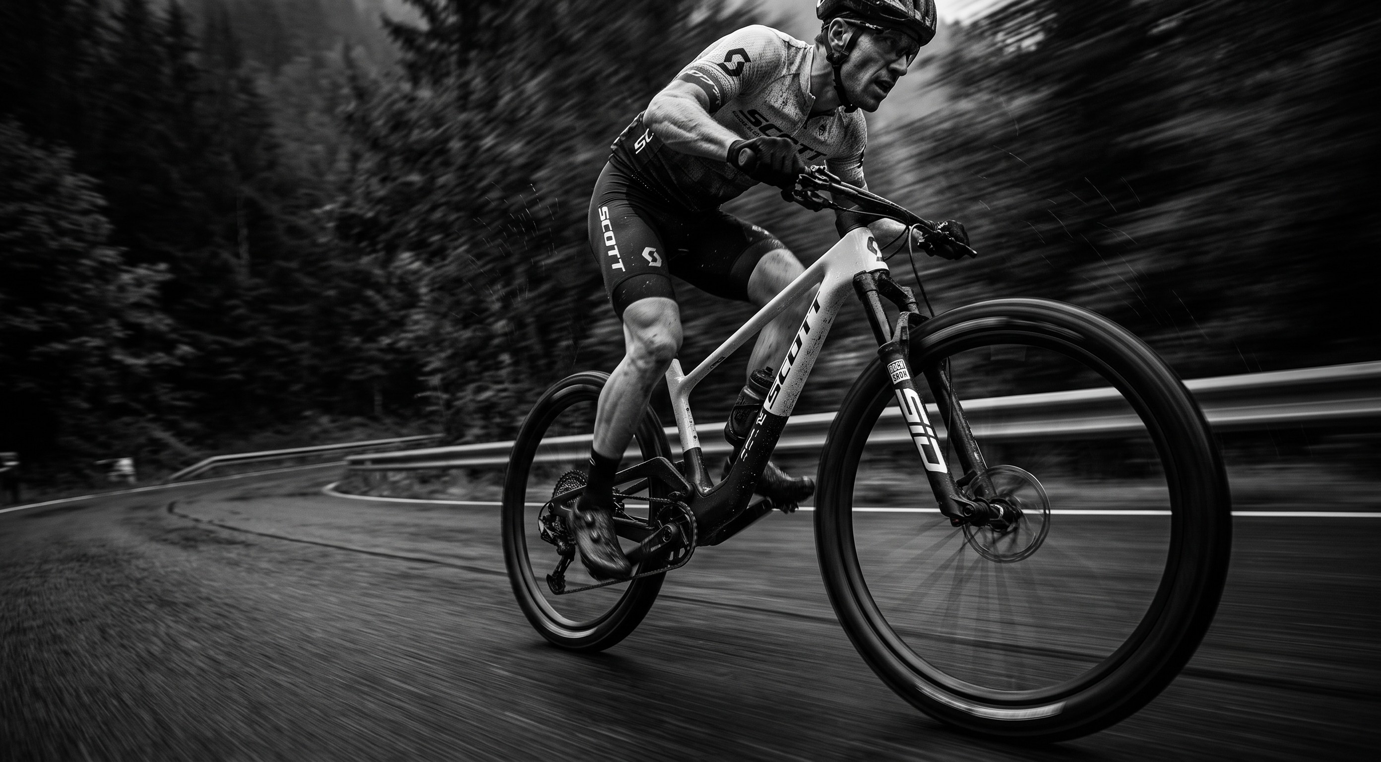 A black and white action shot of a cyclist riding a full-suspension mountain bike on a winding road surrounded by blurred trees. The bike frame and fork show the GORUCKI brand.