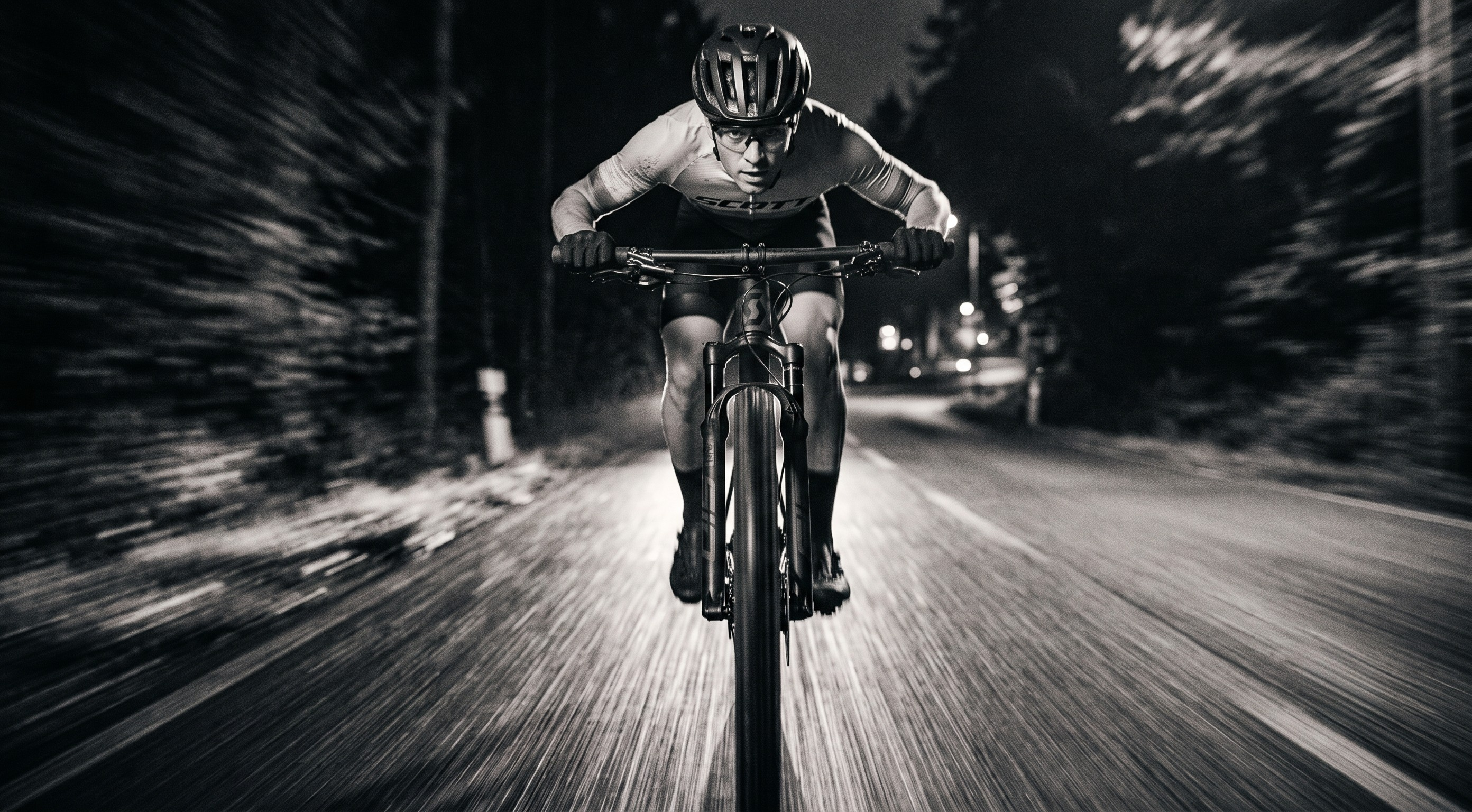 A black and white action shot of a cyclist riding a mountain bike down a dark forest road at night, viewed from a low front-facing angle with motion blur in the background.