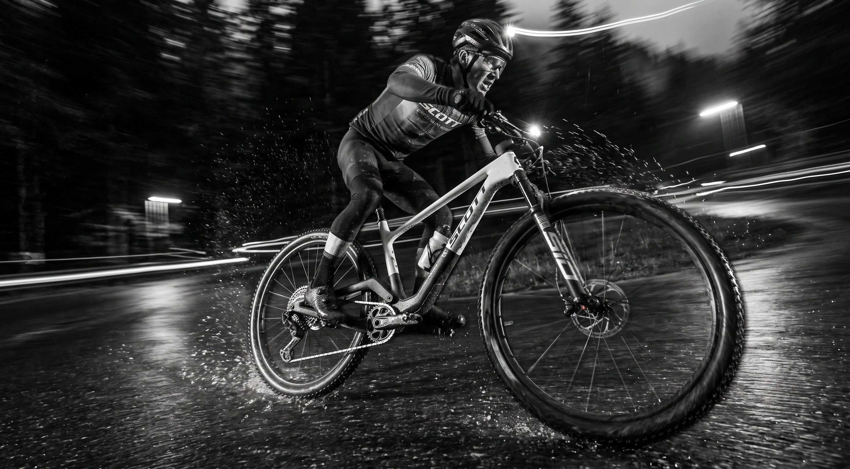 A black and white action photograph of a mountain biker riding a full-suspension bike on a wet road at night, with motion blur and light trails in the background.