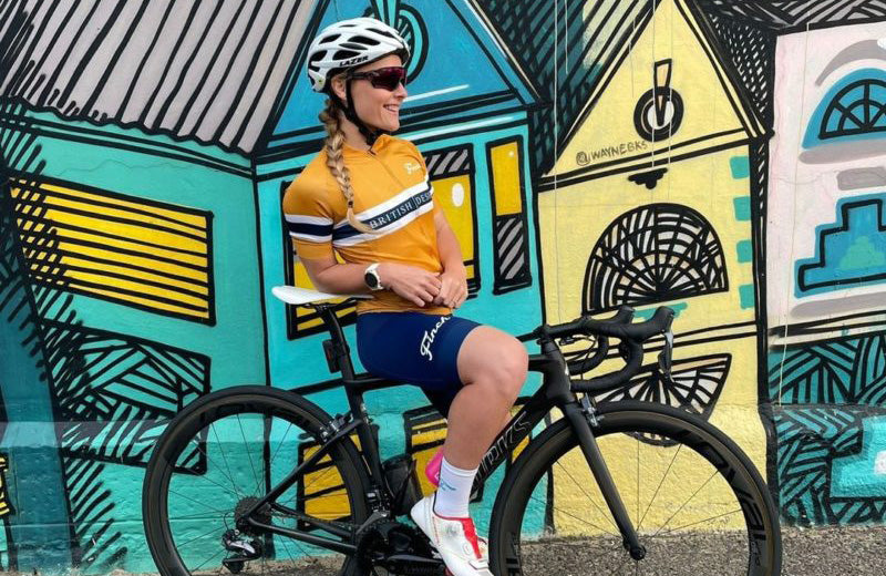 A female cyclist standing still on her bicycle with a painted wall background, the wall features colorful buildings 