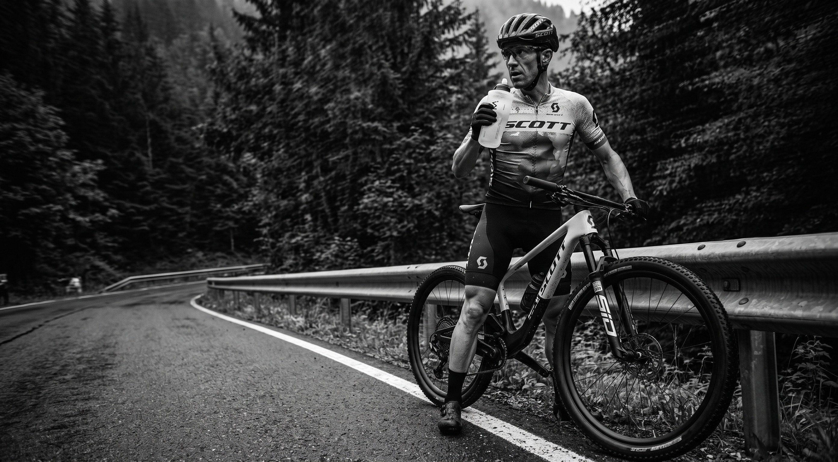 A cyclist wearing a Scott jersey and helmet stands beside a mountain bike on a forest road, holding a bottle. The photo is in black and white, with trees and a guardrail in the background.