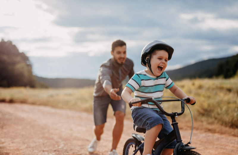 A child riding on a small bike with a male guardian or parental figure standing behind him making sure the child does not fall